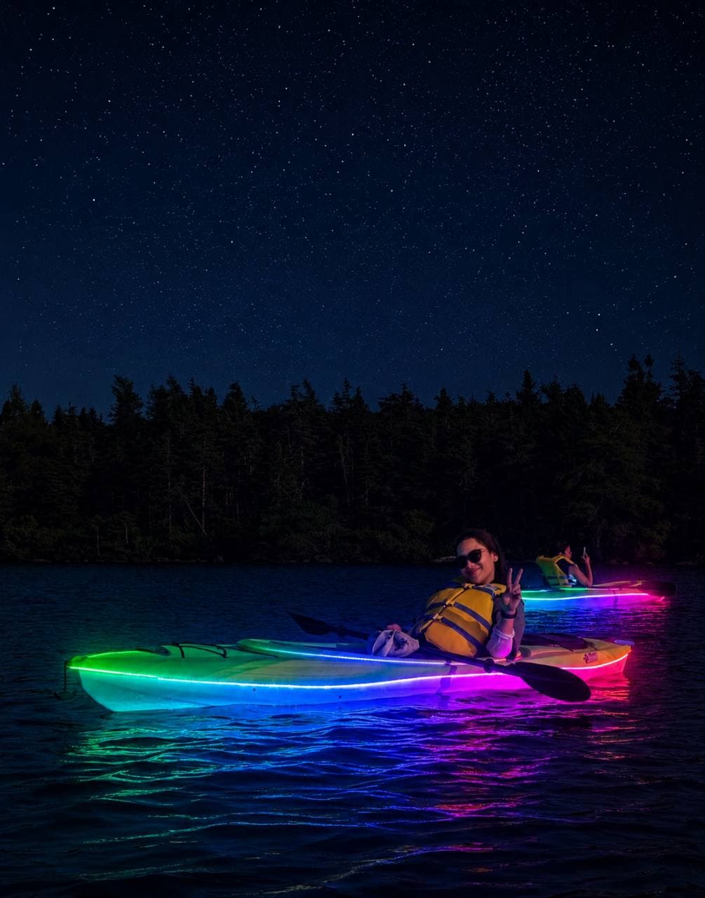 Friends relaxing in RGB LED illuminated kayaks under a star-filled sky.